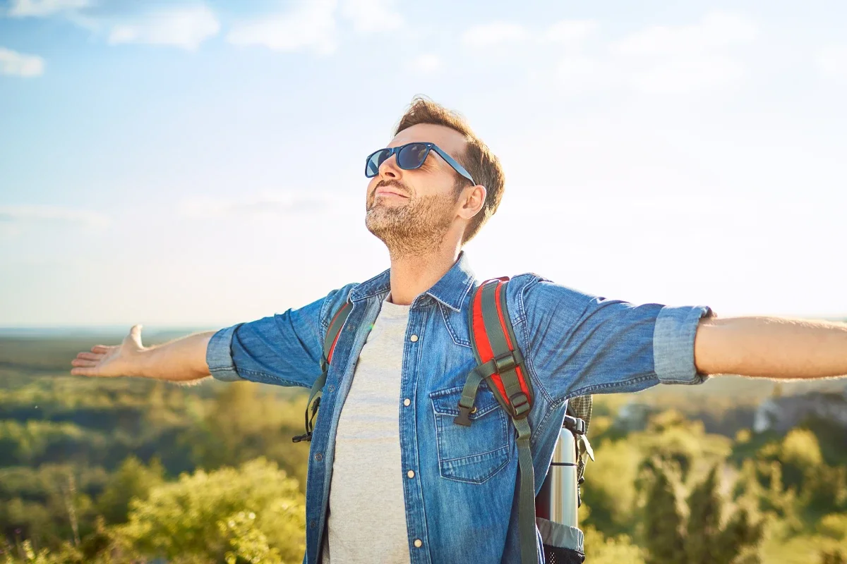 Eine männliche Person mit ausgebreiteten Armen mit Sonnenbrille auf und Rucksack auf